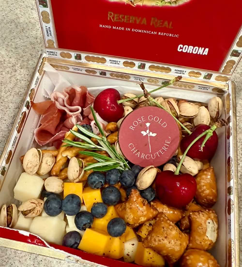 Unique cigar box charcuterie by Rose Gold Charcuterie featuring cubes of cheese, blueberries, cherries, pistachios, pretzels, cured meat, and a branded label inside a Romeo y Julieta cigar box.