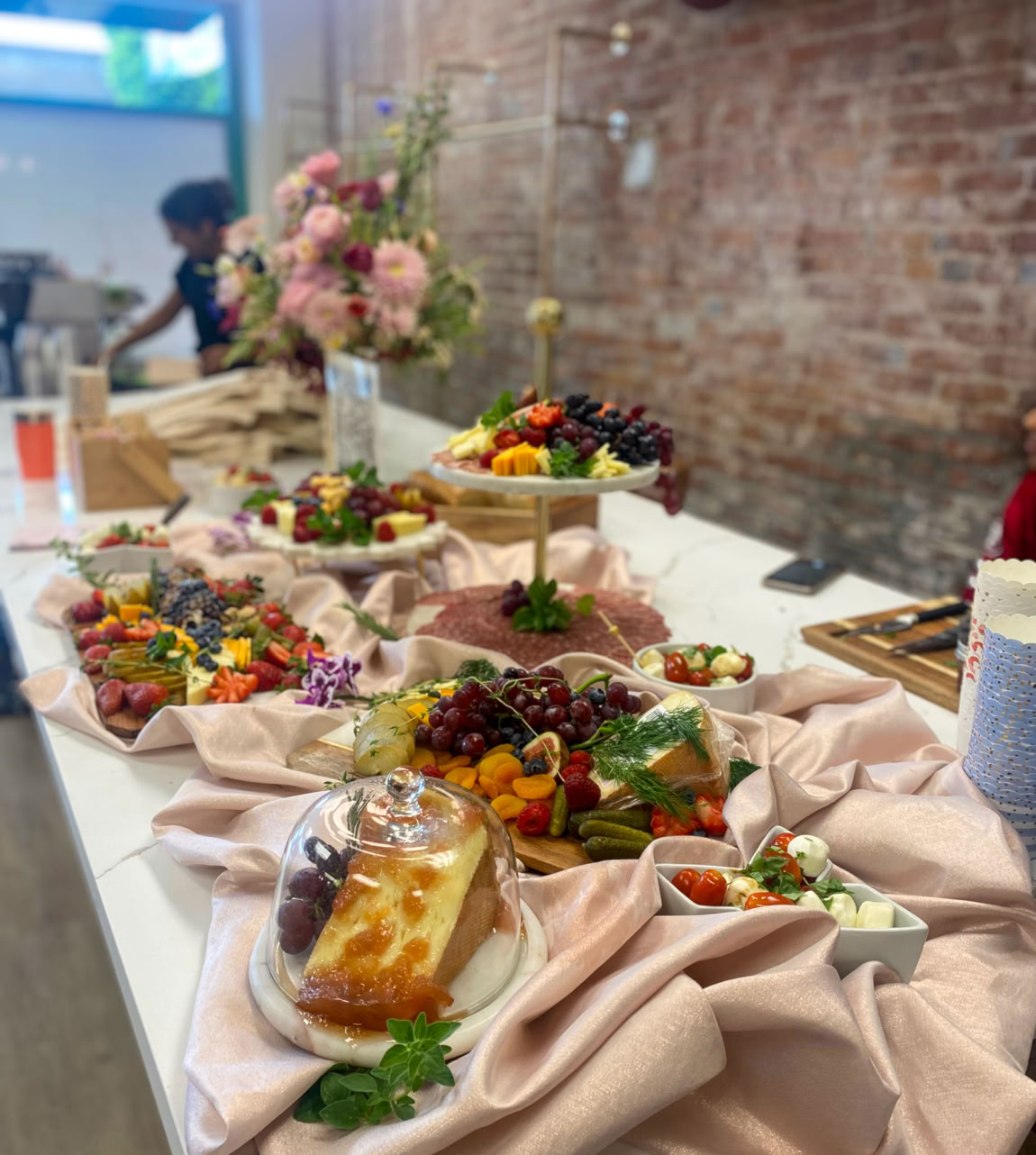 Elegant charcuterie grazing table by Rose Gold Charcuterie featuring gourmet cheeses, fresh fruit, artisanal meats, and seasonal bites styled on blush satin with floral decor and rustic brick backdrop.