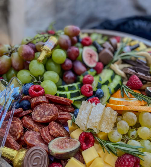 A vibrant charcuterie board featuring an assortment of green and red grapes, sliced cured meats, cubes of cheddar cheese, figs, raspberries, blueberries, honeycomb, orange slices, chocolate pieces, and sprigs of rosemary, all artfully arranged on a wooden platter.