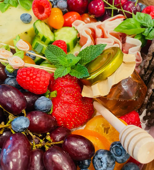 Close-up of gourmet charcuterie box with honey jar, basil, cucumbers, berries, and folded prosciutto.